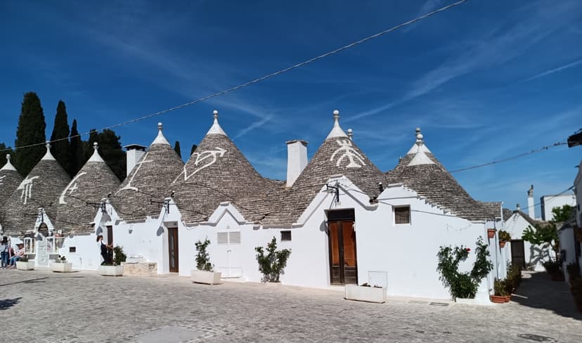 Piedras y trulli: Alberobello y Matera
Alberobello y Matera son dos de los destinos más fascinantes y únicos del sur de Italia, cada uno con una arquitectura característica que los ha convertido en Patrimonio de la Humanidad de la UNESCO.
Alberobello, situada en la región de Puglia, es famosa por sus trulli, construcciones rurales de piedra calcárea con techos cónicos cubiertos de lajas de piedra gris. Estas singulares viviendas, cuyo origen se remonta a la Edad Media, fueron construidas sin argamasa, lo que permitía desmantelarlas rápidamente para evitar el pago de impuestos al reino de Nápoles. El barrio de Rione Monti, con sus más de mil trulli encalados, ofrece un paisaje de cuento de hadas que parece sacado de otro tiempo. Muchos de estos trulli están decorados con símbolos pintados en blanco sobre los techos, de origen pagano, cristiano o esotérico, cuyo significado exacto aún hoy es objeto de debate entre los estudiosos.
Matera, en la región de Basilicata, es conocida por sus Sassi, un complejo sistema de viviendas rupestres excavadas en las laderas de una profunda garganta, el Gravina. Habitados desde el Paleolítico, los Sassi representan uno de los asentamientos humanos continuos más antiguos del mundo. Durante siglos, estas cuevas albergaron a familias enteras que convivían con sus animales en condiciones de extrema pobreza. En la década de 1950, el gobierno italiano ordenó la evacuación forzosa de sus habitantes, considerando los Sassi un símbolo de vergüenza nacional. Sin embargo, a partir de los años ochenta, comenzó un proceso de recuperación y valorización que transformó Matera en una ciudad museo. En 2019, fue nombrada Capital Europea de la Cultura, consolidando su renacimiento cultural y turístico.
